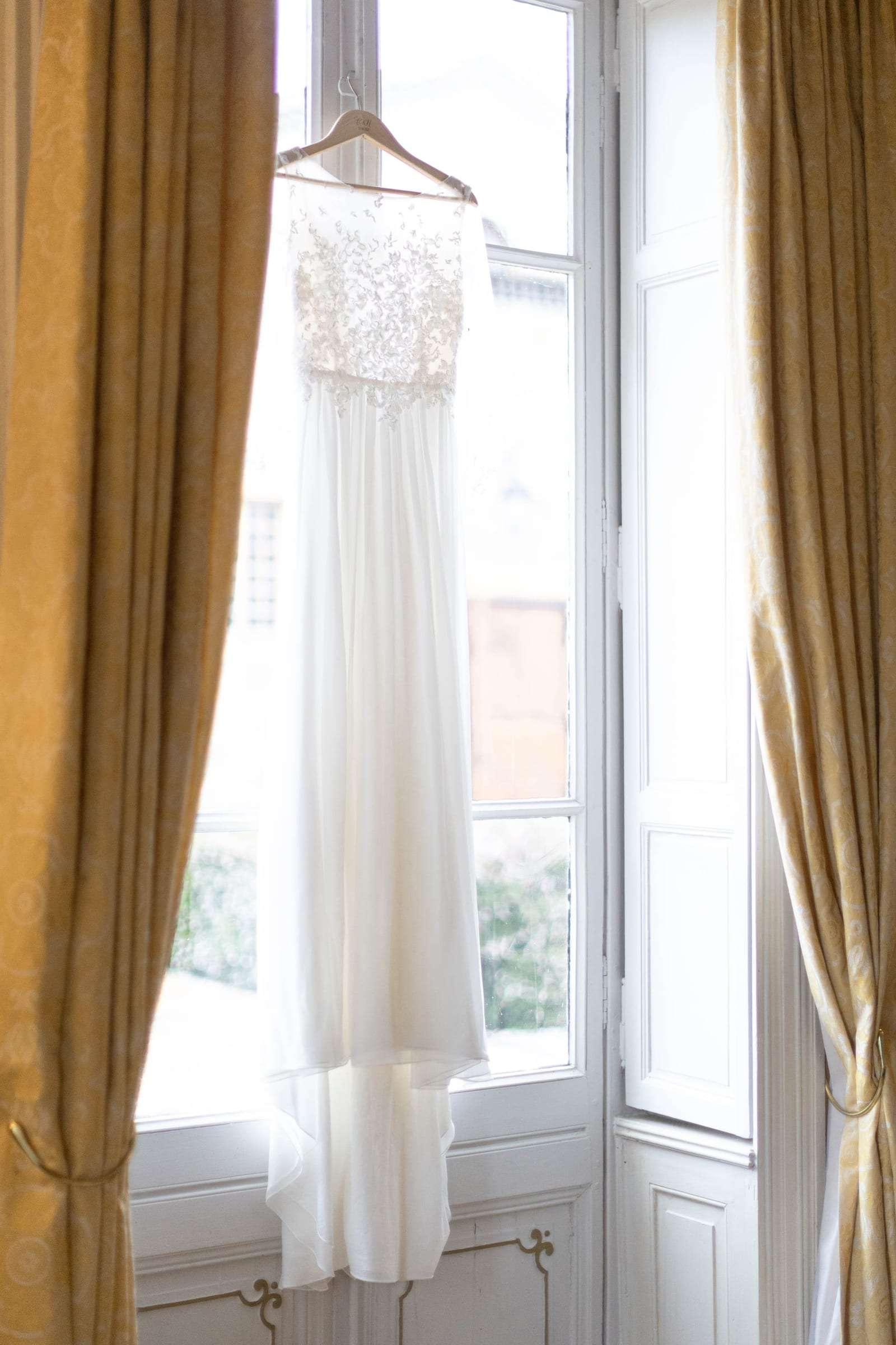 Robe de mariée suspendue devant une grande fenêtre baignée de lumière