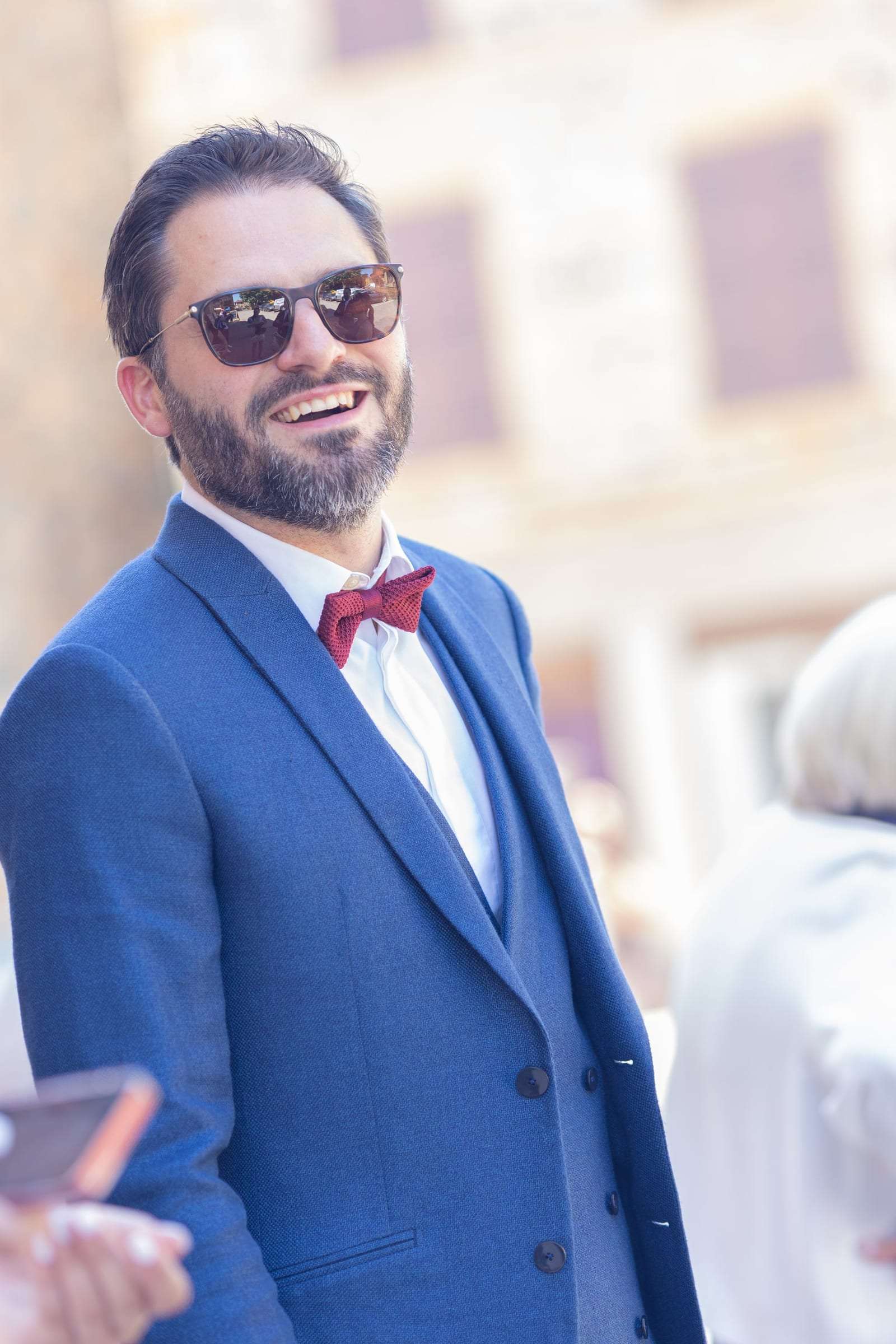 Un invité en costume bleu et nœud papillon rouge sourit derrière ses lunettes de soleil