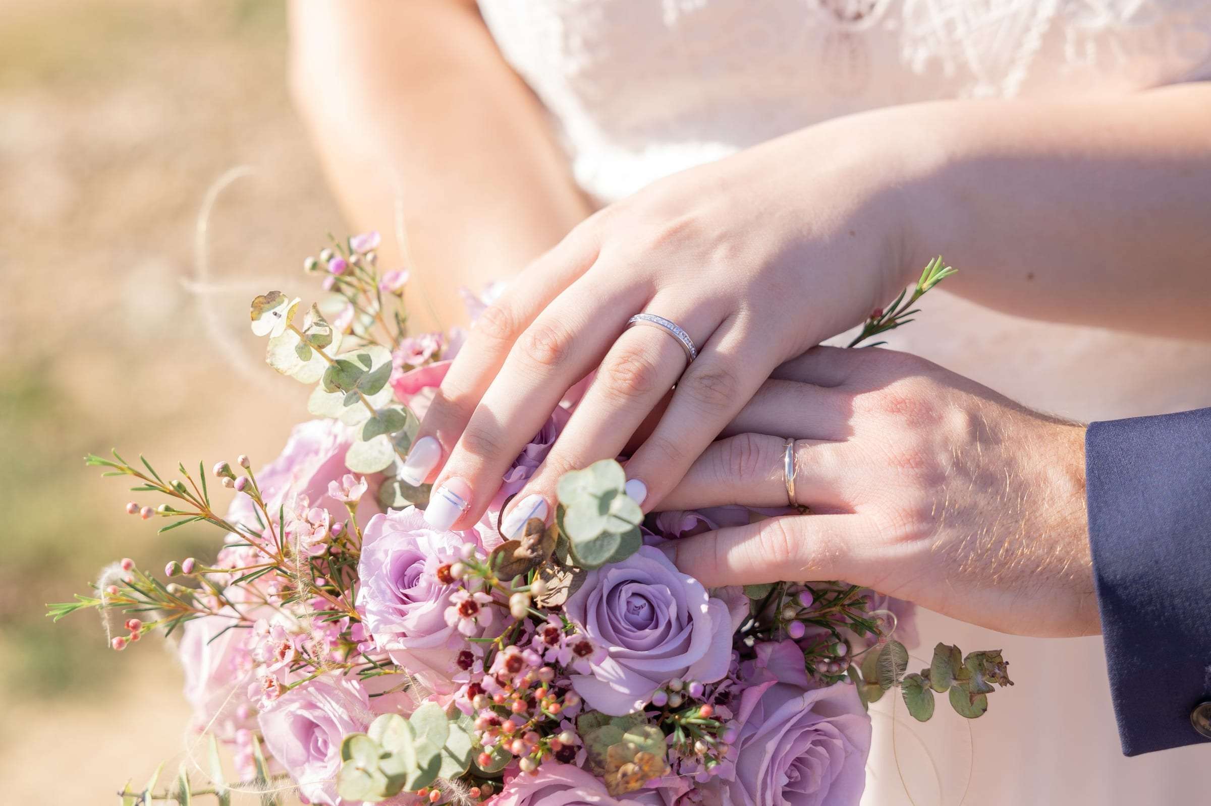 Mains des mariés et alliances posées sur un bouquet aux tons lilas